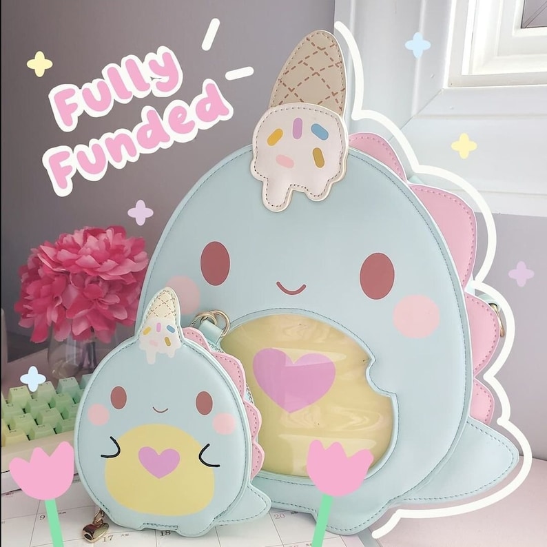 May include: A light blue narwhal-shaped purse with a pink heart and a yellow ice cream cone on top. The purse has a clear window on the front and a smaller matching narwhal coin purse attached to it. The text "Fully Funded!" is written in pink on a light gray background.