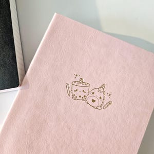 May include: A pink journal with a gold embossed design featuring a cartoon boba tea and a unicorn bear. The journal is closed and has a textured cover. A white box is partially visible in the upper left corner.