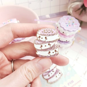 May include: A stack of three enamel pins shaped like cookies with white frosting and purple sprinkles. The pins are gold-toned and have cute cartoon faces on them. The text "ENAMEL PIN" is visible in the image.