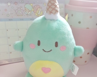 May include: A light blue plush toy shaped like a narwhal with a yellow belly and a pink heart. The narwhal has a white and brown ice cream cone on its head.