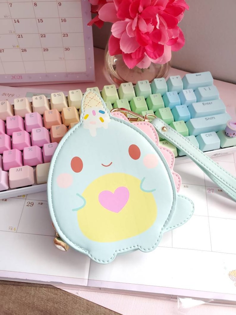 May include: A light blue coin purse with a cartoon whale design. The whale has a yellow belly, a pink heart, and a blue and white ice cream cone on its head. The purse has a gold zipper and a light blue strap.