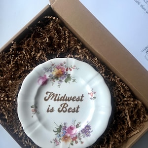 May include: A white decorative plate with floral designs and the text "Midwest is Best" in brown lettering. The plate is inside a cardboard box with brown paper filler.