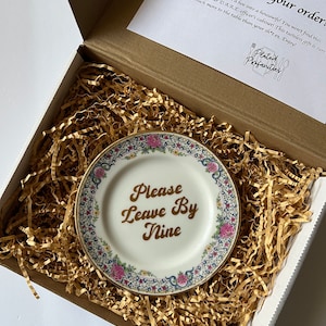 May include: A white porcelain plate with a gold rim and floral pattern. The plate has the words "Please Leave By Nine" written in gold lettering.