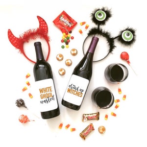 May include: Two wine bottles with Halloween-themed labels. The labels are white with orange text. The left bottle says "White Ghoul Wasted" and the right bottle says "Drink Up Witches". The bottles are surrounded by candy, including Skittles, Twix, and candy corn.