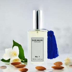 Może przedstawiać: Prostokątny flakon perfum ze srebrną nakrętką i niebieskim chwostem. Etykieta głosi "SUKIMUKI PERFUMES No. 1 100 ml - 3.4 fl. oz." Flakon otoczony jest migdałami, białymi kwiatami i zielonymi liśćmi.