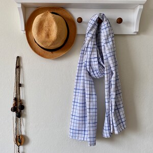 May include: A white and blue checked scarf hangs on a white wooden rack with a straw hat and a beaded necklace hanging next to it.