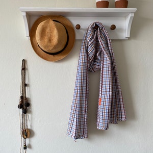 May include: A white shelf with three hooks, a straw hat, a plaid scarf, and a beaded necklace hanging from it. Two small white ceramic cups with brown accents are on the shelf.