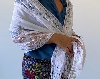 May include: A white lace shawl with a fringed edge, draped over a denim shirt and a floral skirt. The shawl's lace design includes floral and geometric patterns. The overall look is delicate and elegant, suitable for various occasions.