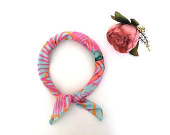 May include: A blue and pink patterned bandana with orange accents. The bandana is tied in a knot and is lying on a white surface.