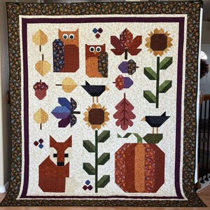 May include: A large, handmade quilt with an autumn theme. The quilt features appliqued images of owls, leaves, sunflowers, a fox, a pumpkin, and acorns in shades of orange, brown, and green on a cream background. The quilt is bordered with a floral pattern.