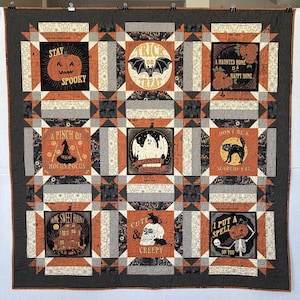 May include: A Halloween-themed quilt featuring orange, black, and cream squares with spooky illustrations and text. Designs include pumpkins, bats, ghosts, cats, and skulls. The quilt has a black and white checkered border.