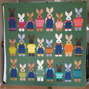 May include: A handmade quilt featuring a repeating pattern of bunny figures. Each bunny is dressed in a different outfit, including overalls and plaid shirts, in various colors. The background is a solid green.