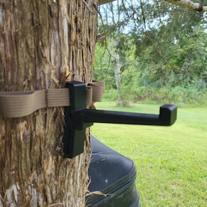May include: A black metal hook mounted to a tree trunk with a brown strap. The hook is designed to hang items from a tree.