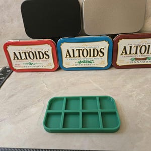 May include: Four Altoids mint tins, one each in red, blue, silver, and black. The tins are arranged in a pyramid shape. The text on the tins reads "The Original Celebrated Altoids Curiously Strong Mints" with the flavor listed below. The flavors are peppermint, wintergreen, cinnamon, and an unreadable flavor. A green plastic tray with nine compartments is in front of the tins.