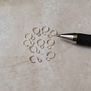 May include: A close-up of a group of silver jump rings, arranged in a circle on a light beige surface. A black pen is lying on the surface, partially visible in the image.