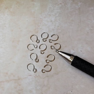 May include: A close-up of a group of silver jump rings on a light gray surface. A black pen is lying on the surface to the right of the jump rings.