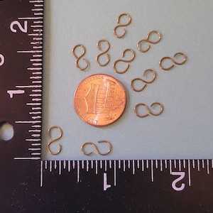 May include: A close-up of nine gold-colored, small, infinity-shaped jump rings. The jump rings are scattered on a light blue surface. A penny is in the center of the image for size comparison.