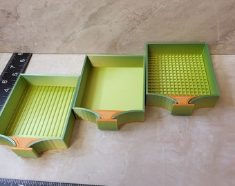 May include: Three-tiered, 3D-printed storage trays in shades of green and orange. The trays have varying interior designs, including ribbed and grid patterns. The trays are arranged in a stepped configuration, with a ruler in the foreground.