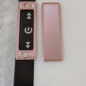 May include: A light pink rectangular device with a black screen displaying a power symbol and up/down arrows. Two silver magnetic circles are on the device. A separate light pink rectangular piece is next to it.