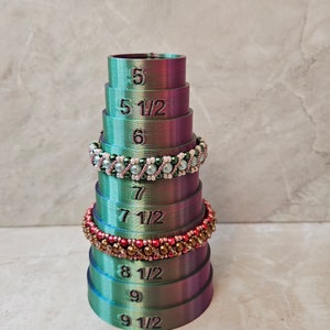 May include: A stack of 10 green and purple ring sizers with numbers from 5 to 9 1/2. The sizers are made of plastic and have a smooth finish.