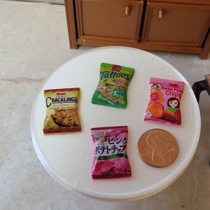 May include: Four miniature bags of chips on a white surface. The bags are labeled "Cracklings", "Tattoos Corn Chips", "Sweet Potato Chips", and "ビンダッチポテトチップス". A copper-colored US penny is also on the surface.