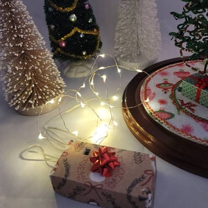 May include: A festive Christmas scene featuring miniature decorated trees and a string of warm white fairy lights. A small, gift-wrapped box with a red bow sits in the foreground. The box is wrapped in brown paper with a holiday-themed pattern.