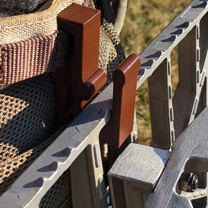 May include: Two brown plastic hooks attached to a metal frame. The hooks are designed to be used with a backpack or other gear.