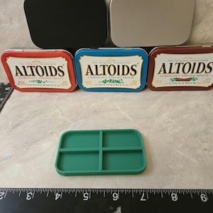 May include: Four empty Altoids mint tins, two silver, one black, and one red. The tins are arranged in a row. Below the tins is a green plastic tray with four compartments.
