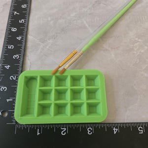 May include: A bright green paint palette with 15 square wells and a handle, paired with two paint brushes. The brushes have light green and clear handles with gold and tan bristles. A black ruler is next to the palette.