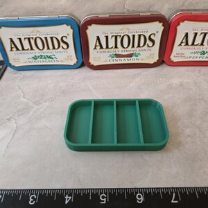 May include: A green rectangular organizer with four compartments sits in front of three Altoids tins. The tins are blue, maroon, and red, with the Altoids logo and flavor names visible: Wintergreen, Cinnamon, and Peppermint.