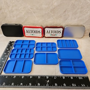 May include: Six blue plastic containers with different compartment configurations. The containers are arranged in two rows of three. The containers are sitting on a black ruler with white markings. The ruler is marked in inches. Two red and silver Altoids tins are visible in the background.