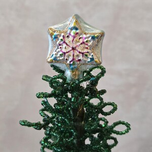 May include: A Christmas tree topper with a star-shaped ornament. The star is silver with gold trim and a colorful snowflake design. The tree is made of green beads.