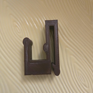 May include: A dark brown, U-shaped plastic clip with a rectangular opening and a rounded end. The clip has a textured surface and appears to be 3D-printed. The clip is designed to hold something in place.