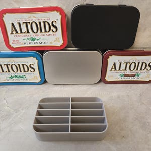 May include: An assortment of Altoids mint tins in red, blue, and brown, with the text "Altoids" and flavor names. A black tin and a silver tin are also present. A gray plastic organizer with multiple compartments is in the foreground.