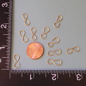 May include: A close-up of nine gold-colored metal wire loops, resembling the number eight, scattered on a light blue surface. A US penny is included for scale.