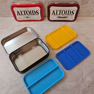 May include: Two empty Altoids tins with the original peppermint and cinnamon flavors. The tins are open and show the inside of the tins. There are three 3D printed inserts for the tins, one yellow and two blue.