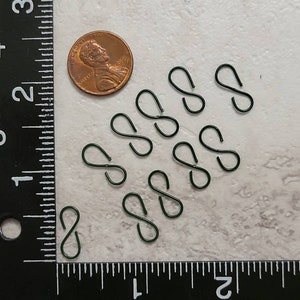 May include: Ten small, black, metal S-shaped hooks, approximately the size of a US penny, are arranged in a semi-circle on a white surface. A ruler is visible in the bottom of the image.