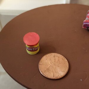 May include: A small red and yellow Play-Doh container next to a US penny dated 2021.