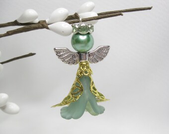 May include: A green glass angel ornament with gold trim and silver wings. The angel is hanging from a branch.