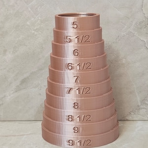 May include: A stack of rose-gold colored rings, each labeled with a size from 5 to 9 1/2. The rings are arranged in a conical shape, with the largest ring at the base and the smallest at the top. The numbers are clearly visible.