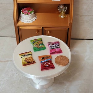 May include: Four miniature bags of chips on a white cake stand. The bags are labeled "Cracklings", "Tattoos", "Cheetos Party Size!" and "ピスタチオ (Pistachio)". A penny is also on the stand.