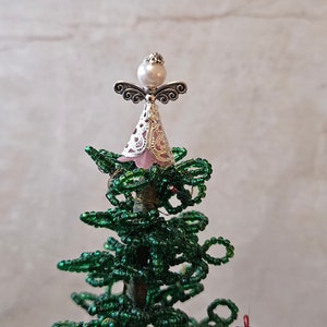 May include: A small, green beaded Christmas tree with a pink and silver angel topper. The angel has a pearl in the center of its head.
