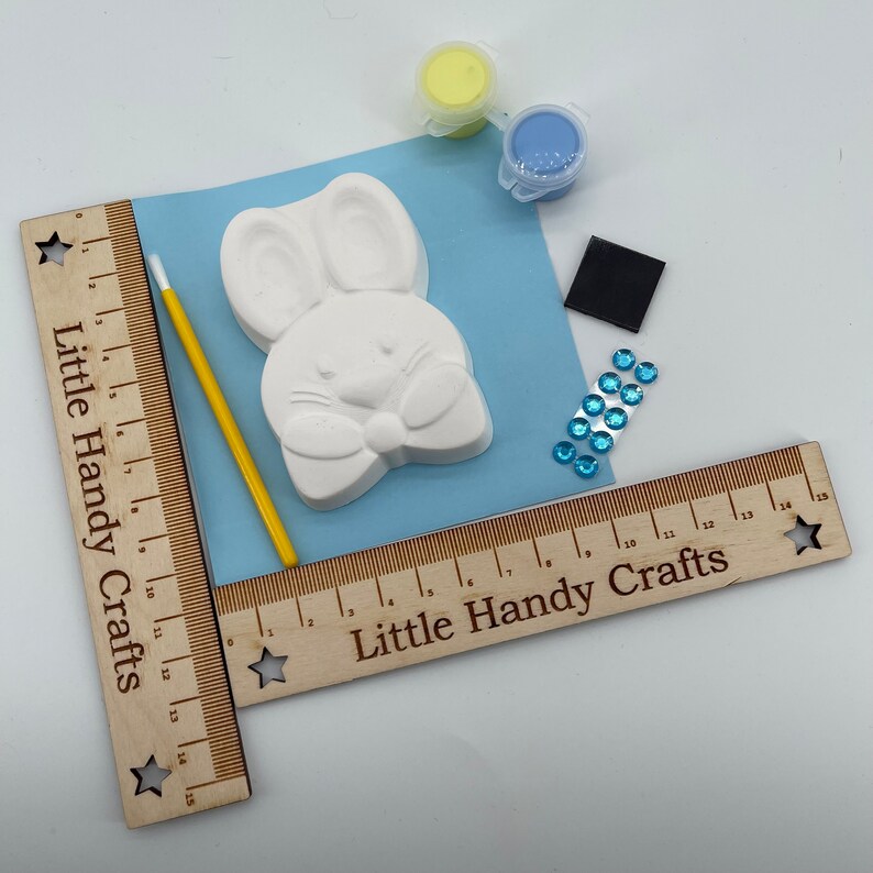 May include: A craft kit featuring a white bunny figure, paint, a paintbrush, and embellishments. The kit includes yellow and blue paint, a black square, and blue rhinestones. The text "Little Handy Crafts" is printed on the wooden ruler.
