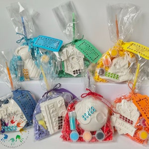 May include: A collection of craft kits in clear cellophane bags. Each bag contains a plaster figure, paint pots, a paintbrush, and decorative elements. The bags are tied with ribbons and include small tags with handwritten text. The website address littlehandycrafts.co.uk is visible.
