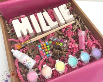 May include: A craft kit in a cardboard box with the name "MILLIE" in white block letters. The kit includes paint, glitter, a paintbrush, glue, and colorful gems, all arranged on pink paper shreds.