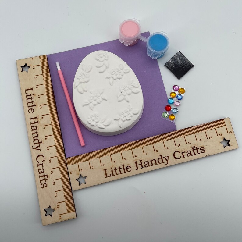 May include: A craft kit with a white egg-shaped plaster mold decorated with floral designs. The kit includes paint, a paintbrush, gems, and a sanding block. Wooden rulers with the text "Little Handy Crafts" frame the scene.