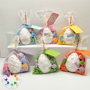May include: Six Easter egg craft kits, each in a clear cellophane bag tied with a ribbon and a tag. The kits include paint, glitter, and a ceramic egg. The bags are filled with colourful paper shreds.