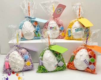 May include: Six Easter egg-shaped craft kits, each in a clear cellophane bag tied with a ribbon and a tag. The kits include paint, glitter, and a ceramic egg. The bags are filled with colorful paper shreds.