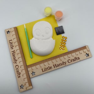 May include: A craft kit with a ceramic chick hatching from an egg, paint pots, a paintbrush, and decorative gems. The kit includes a wooden ruler with the text "Little Handy Crafts". The chick is on a yellow surface.