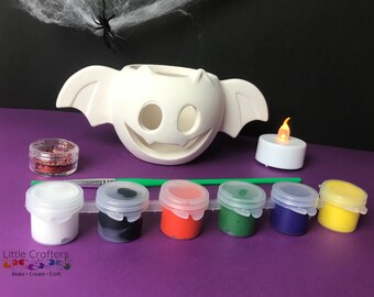 May include: A white ceramic bat-shaped candle holder with wings, a small jar of red glitter, five paint pots in white, black, red, green, and blue, a green paintbrush, and a white tea light with a flickering flame.